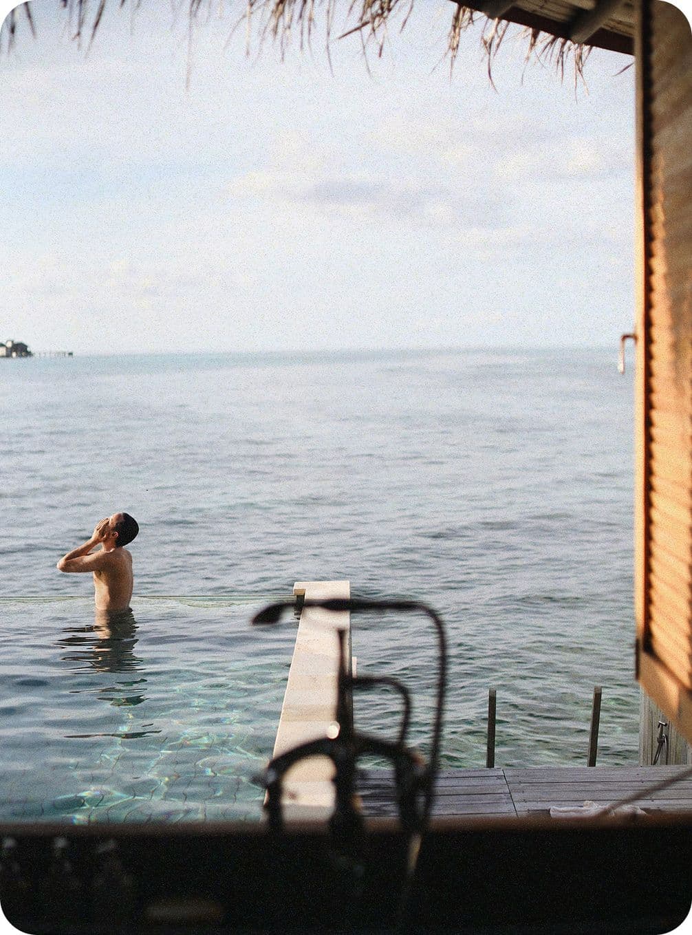 A person stands in an infinity pool overlooking a calm ocean, with a wooden deck and thatched roof visible in the foreground.