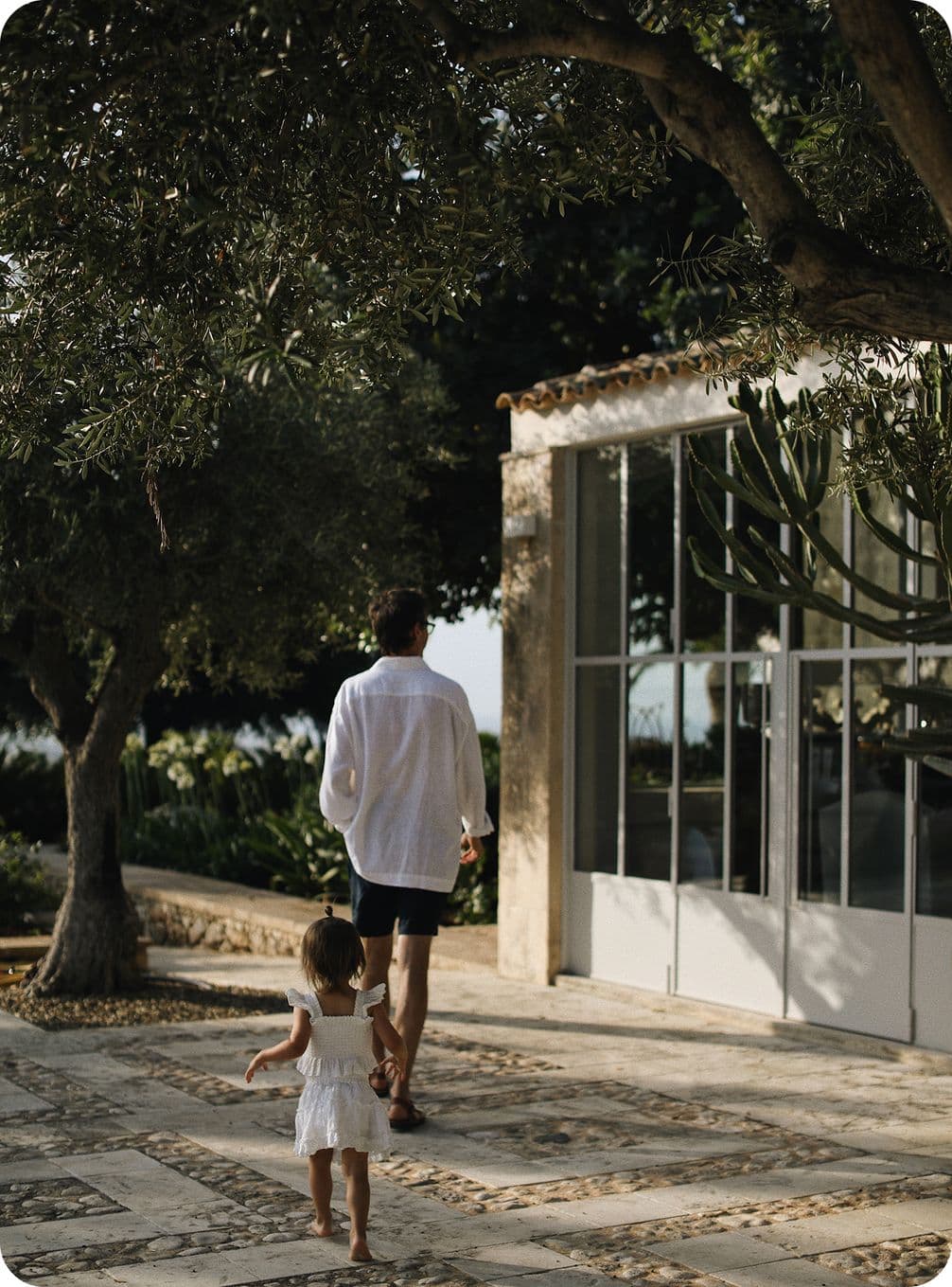 Man and child walking on a stone path beside a glass-walled building, surrounded by trees and greenery.