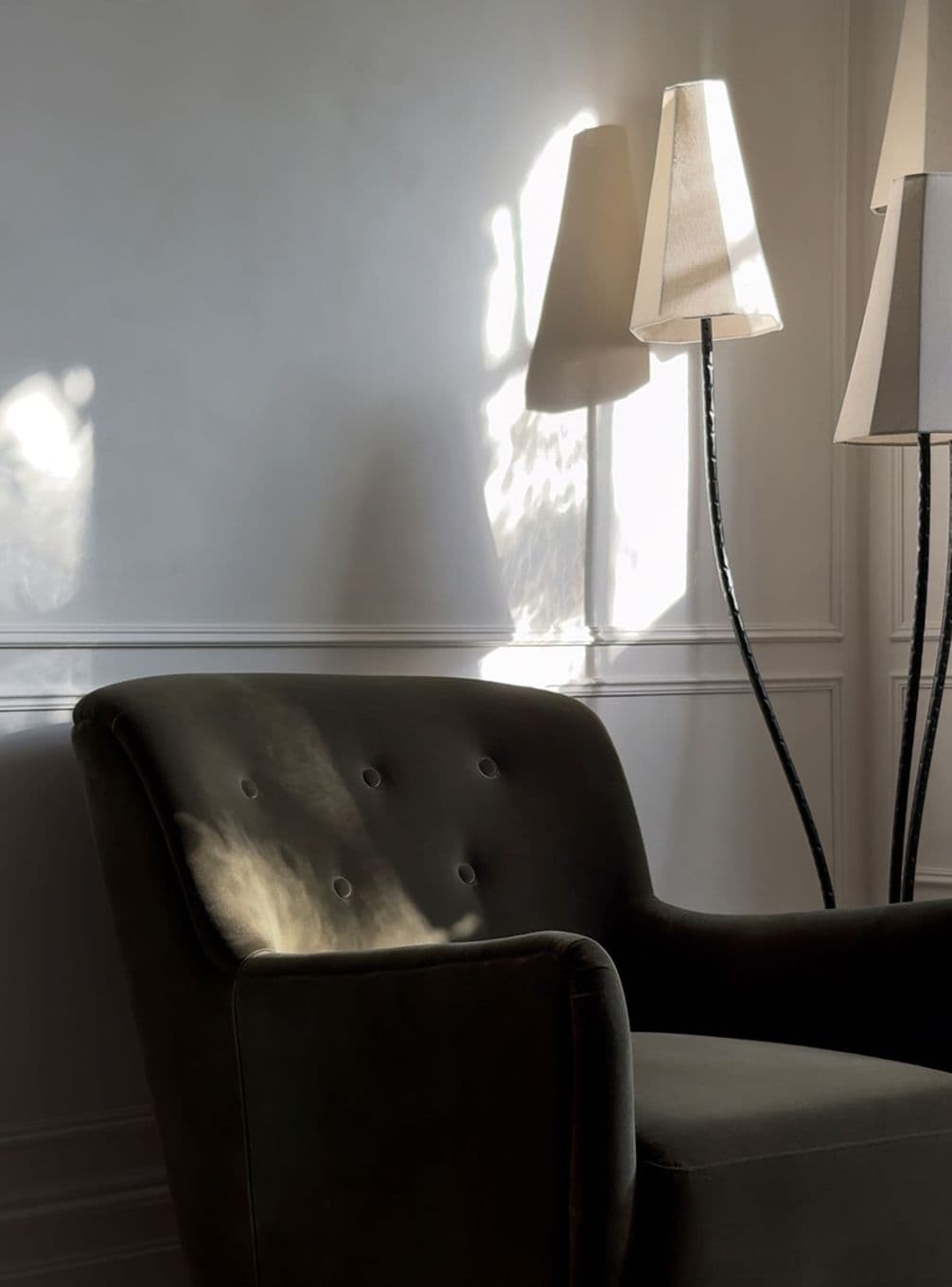 A shadowed, dark armchair with angular lamps casts soft light and shadows on a white paneled wall. Minimalist and serene interior setting.