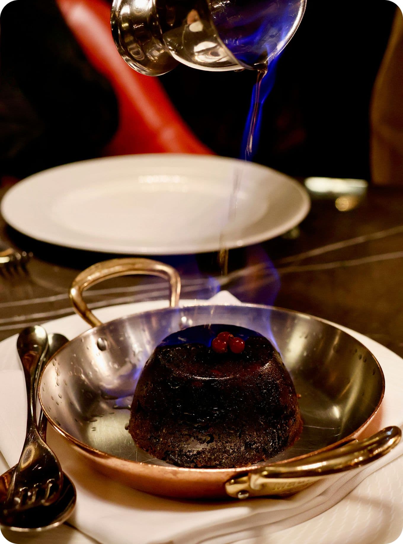 A flaming dessert with berries is being flambéed in a metal pan, with a serving spoon nearby on a white plate.