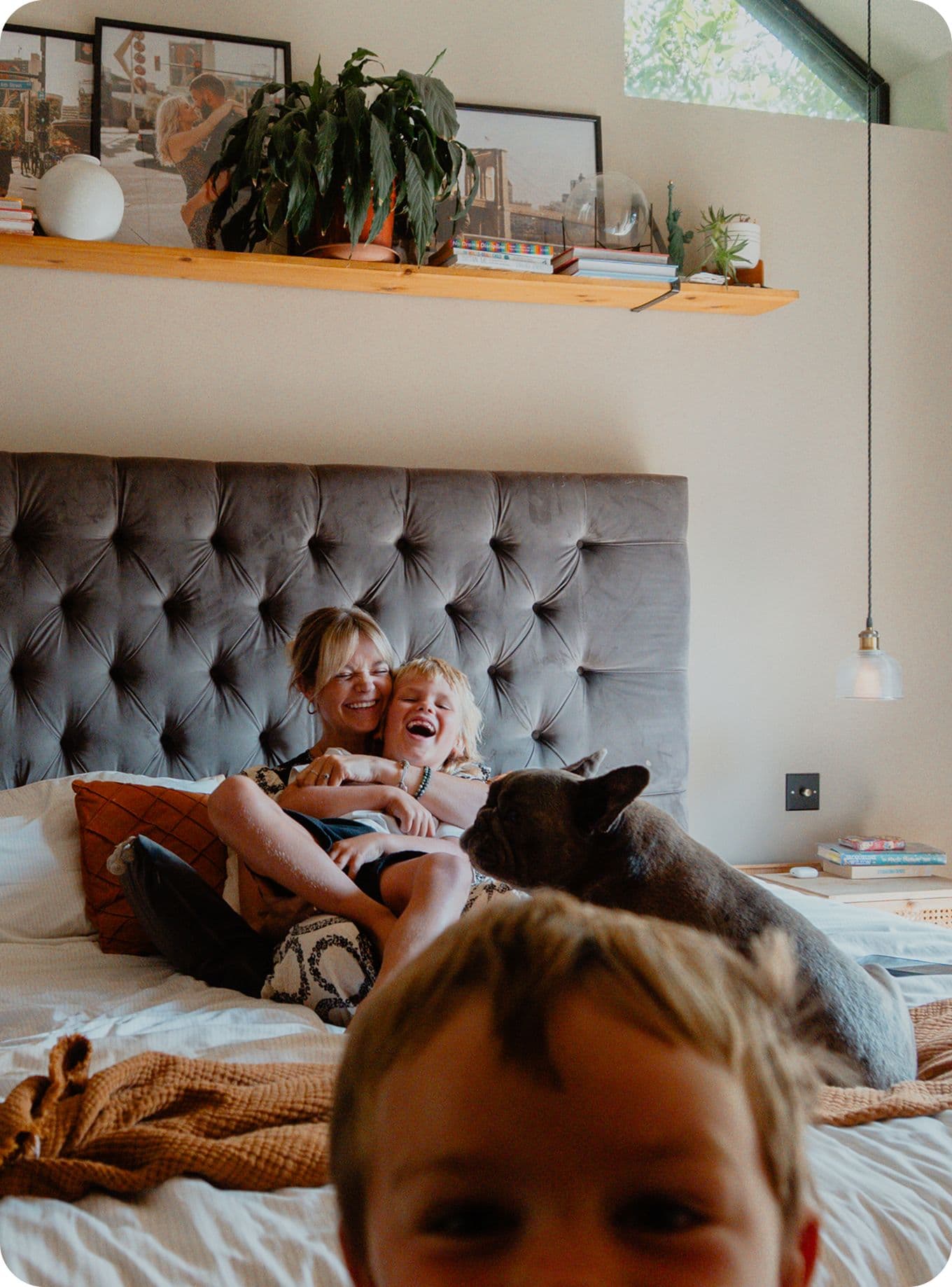 A woman and two children laugh on a bed with a dog. A shelf with plants and photos is above them. Cozy bedroom setting.