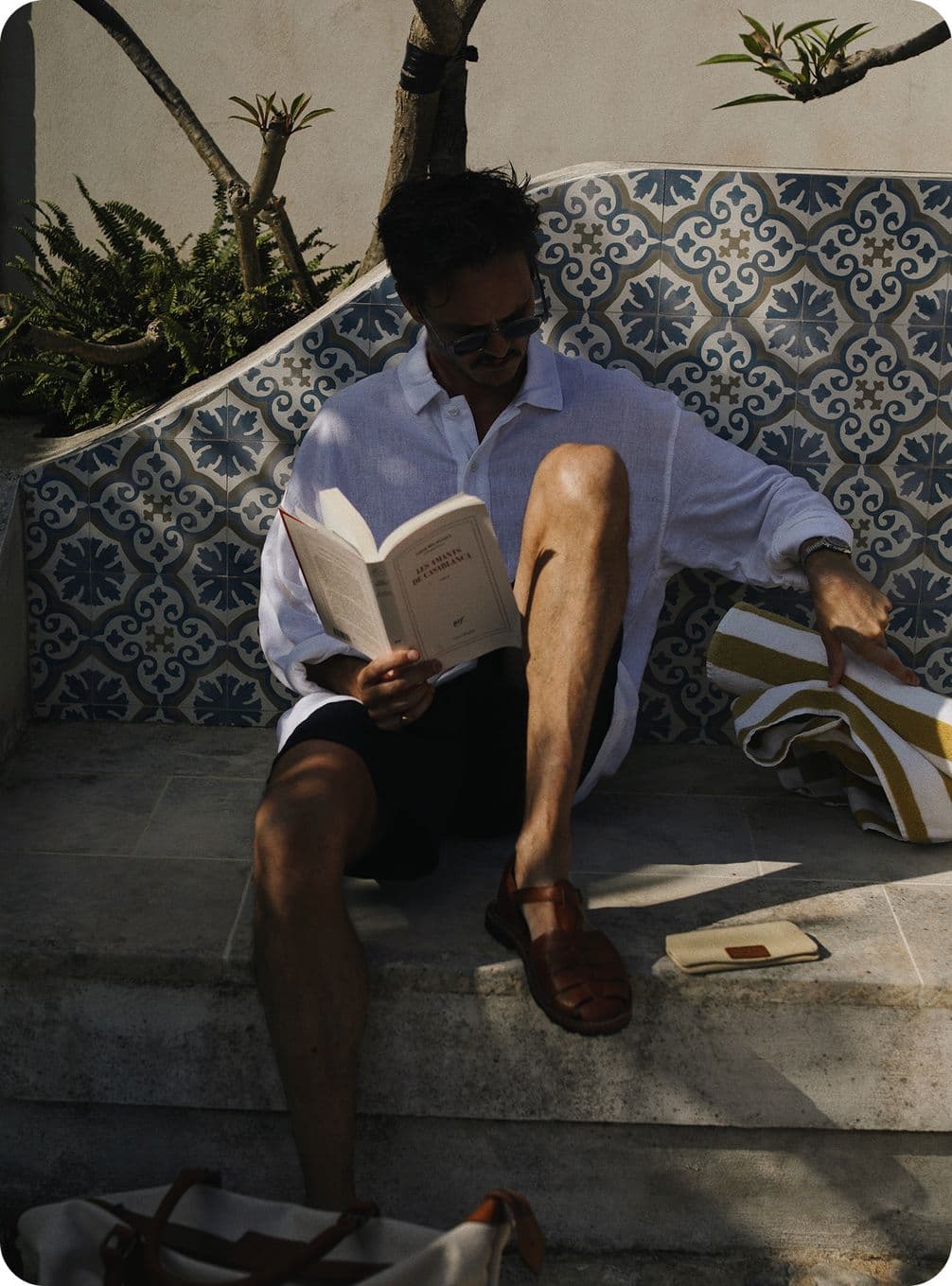 Man in white shirt reading on a patterned bench, with a tote bag beside him, under partial shade.