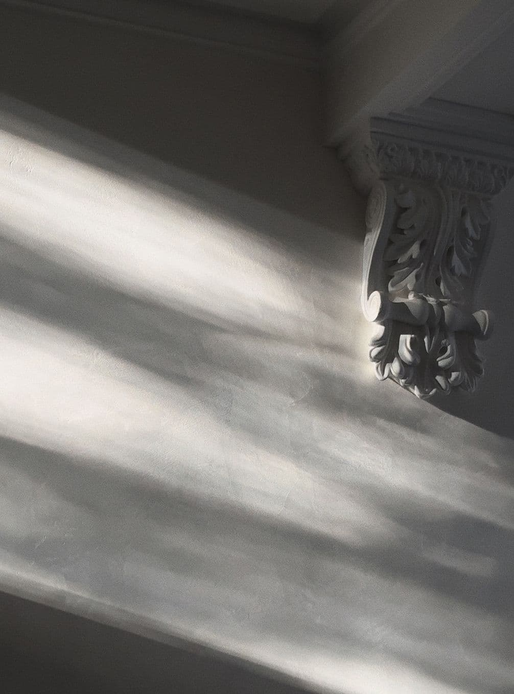 Sunlight casting shadows on a wall with a decorative plaster corbel, creating a soft, serene atmosphere.