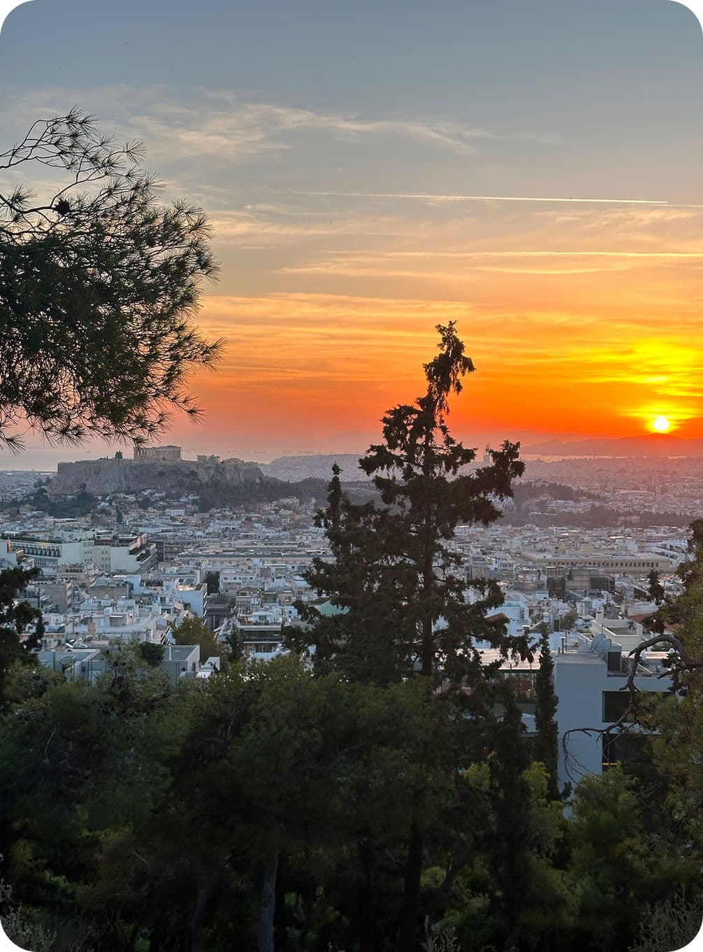 Sunset view over a cityscape with the Acropolis in the distance, framed by silhouetted trees and a vibrant orange sky.