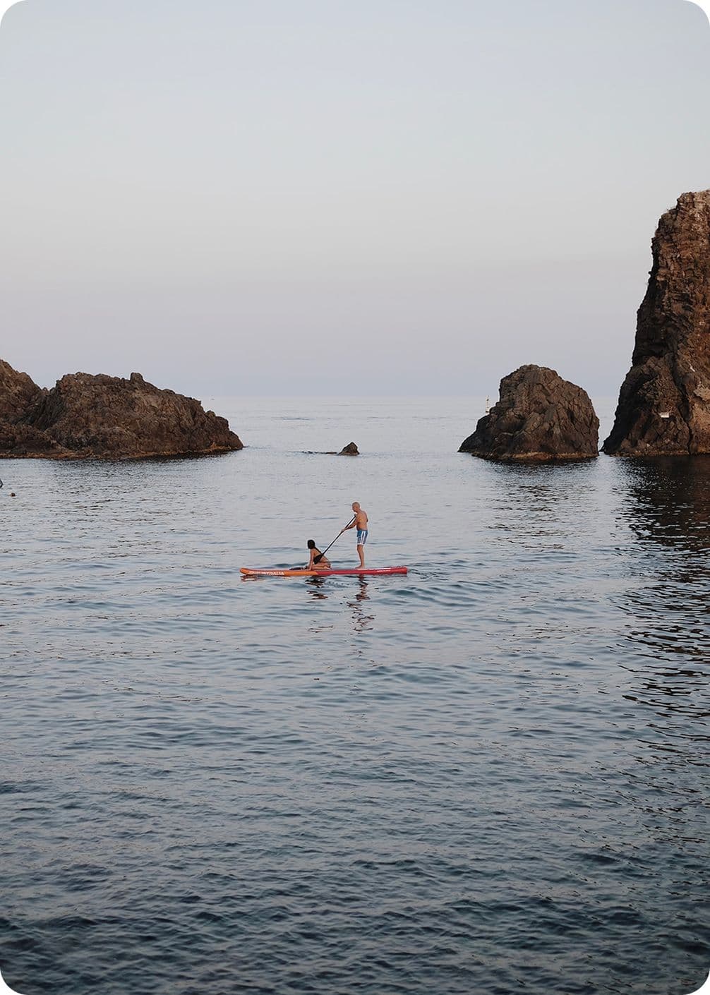 Two people on a paddleboard, one standing and one seated, paddling between rocky outcrops on a calm sea under a pale sky.