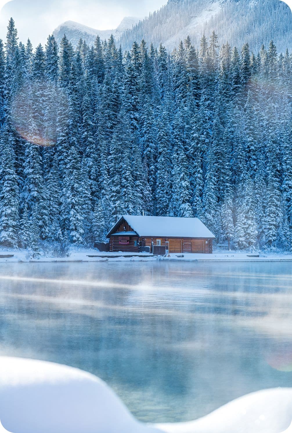 A cozy cabin sits beside a tranquil, misty lake, surrounded by snow-covered pine trees and mountains under a clear blue sky.
