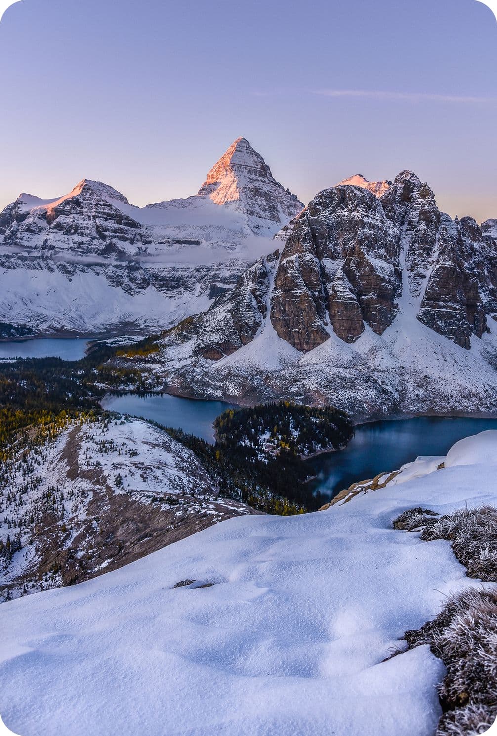 Snow-covered mountains with a prominent peak at sunrise, overlooking a valley with lakes and evergreen trees.