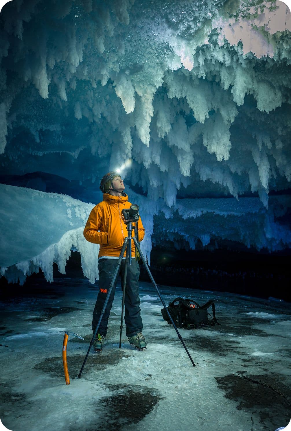 Person in an orange jacket and helmet stands with a tripod in an ice cave, looking up at icicles and formations, with a headlamp illuminating the scene.