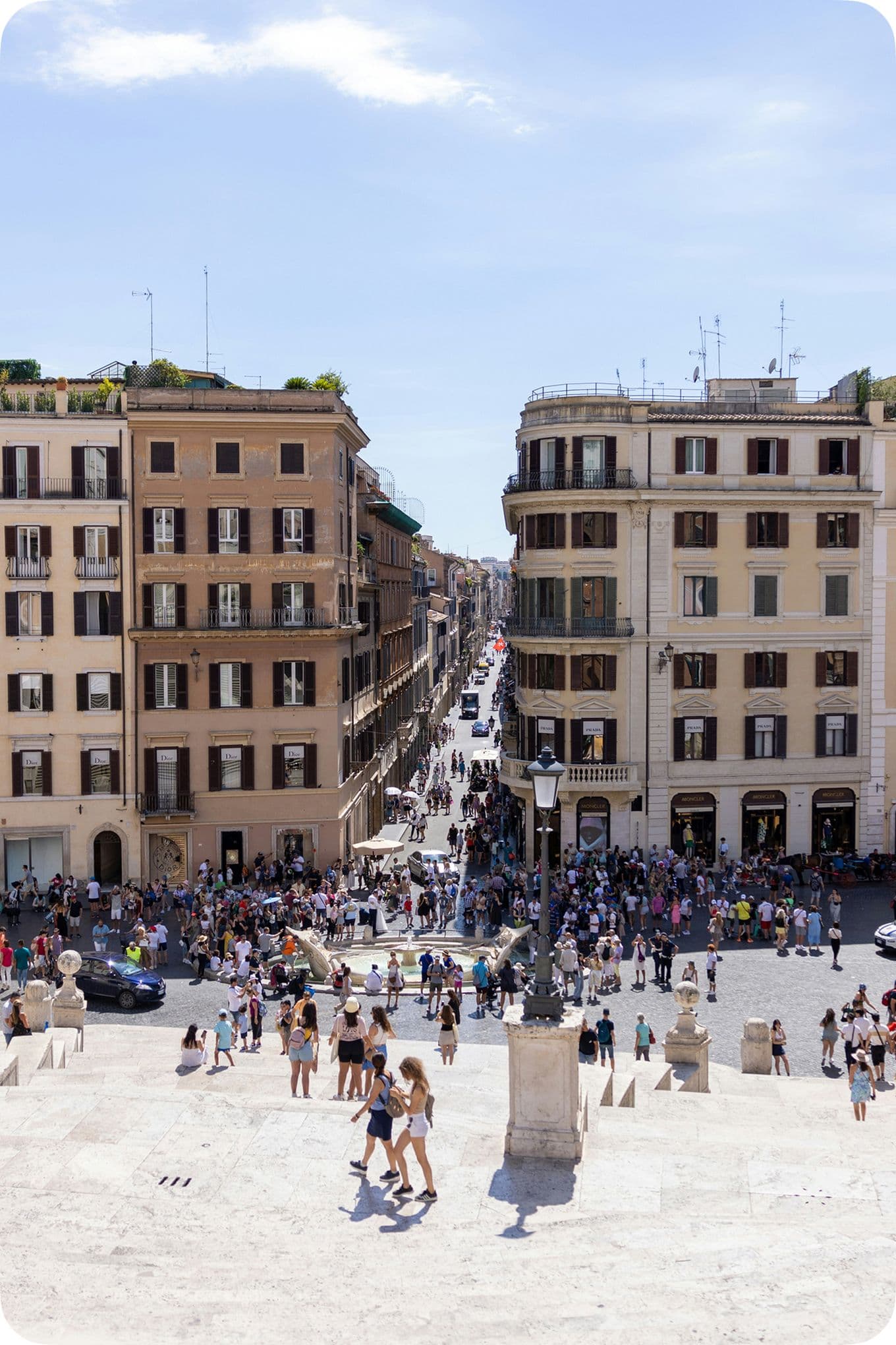 View from Spanish Steps in Rome, overlooking a bustling street with crowds of people and historic buildings under a clear blue sky.