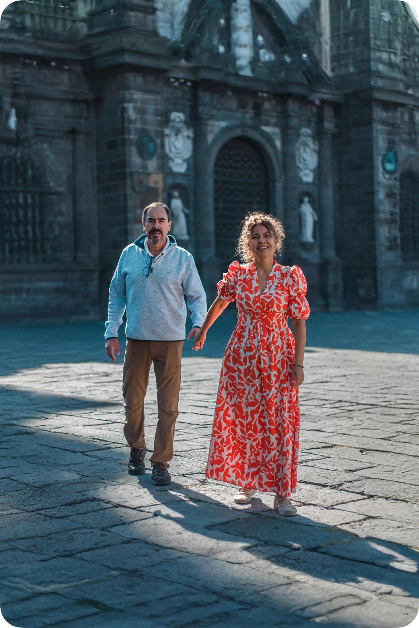 A man and woman hold hands while walking on a cobblestone street, with a historic stone building in the background.