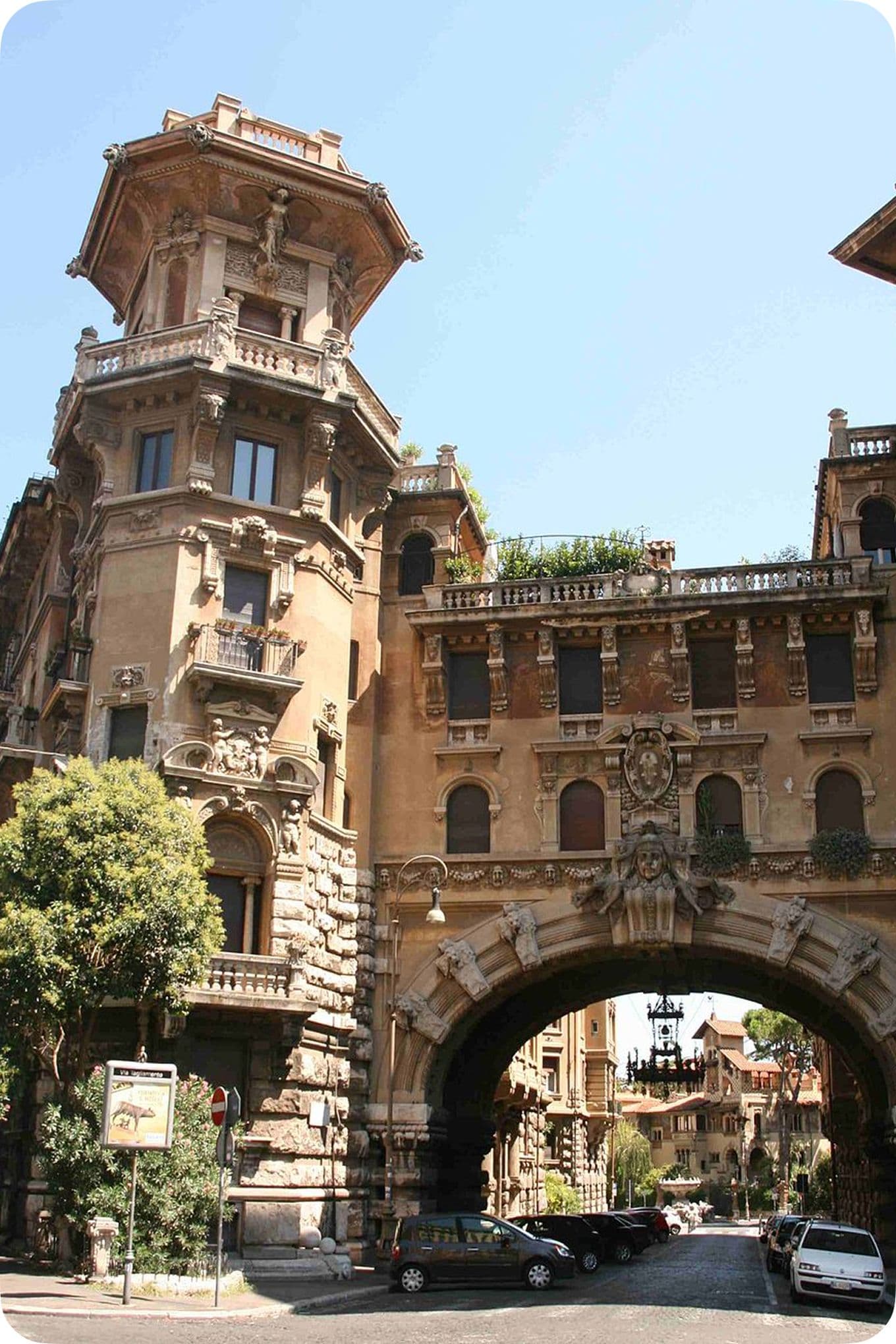 Historic building with ornate architecture featuring an archway over a street, surrounded by trees and parked cars on a sunny day.