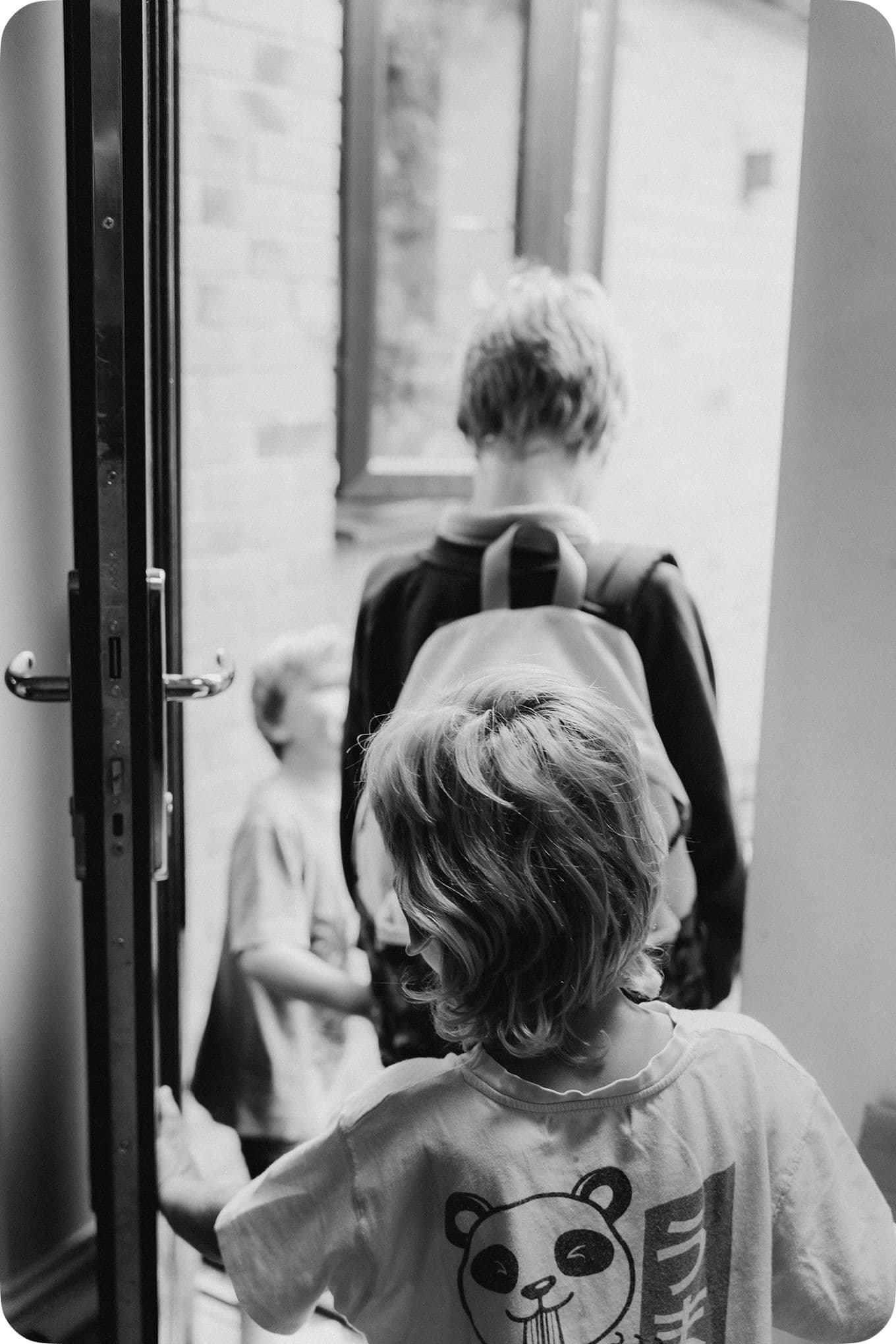 Three children, one with a panda shirt, stand at an open door, looking outside. The scene is in black and white.