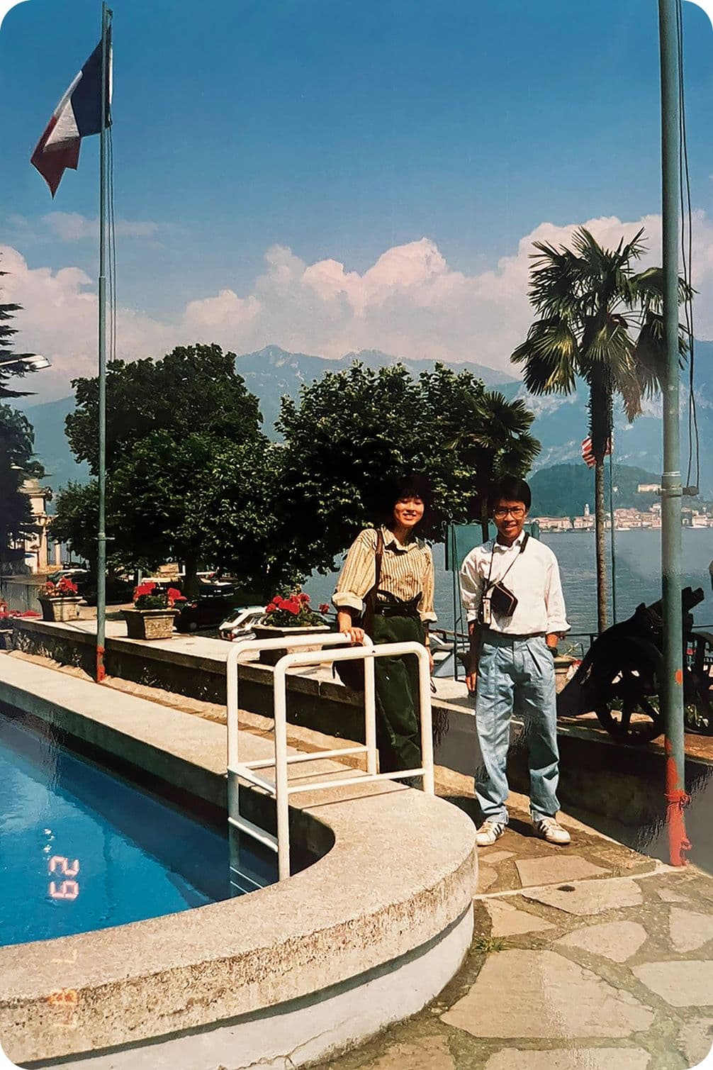 Two people stand by a pool with a scenic backdrop of mountains, palm trees, and a lake. Flags are visible, and the sky is partly cloudy.