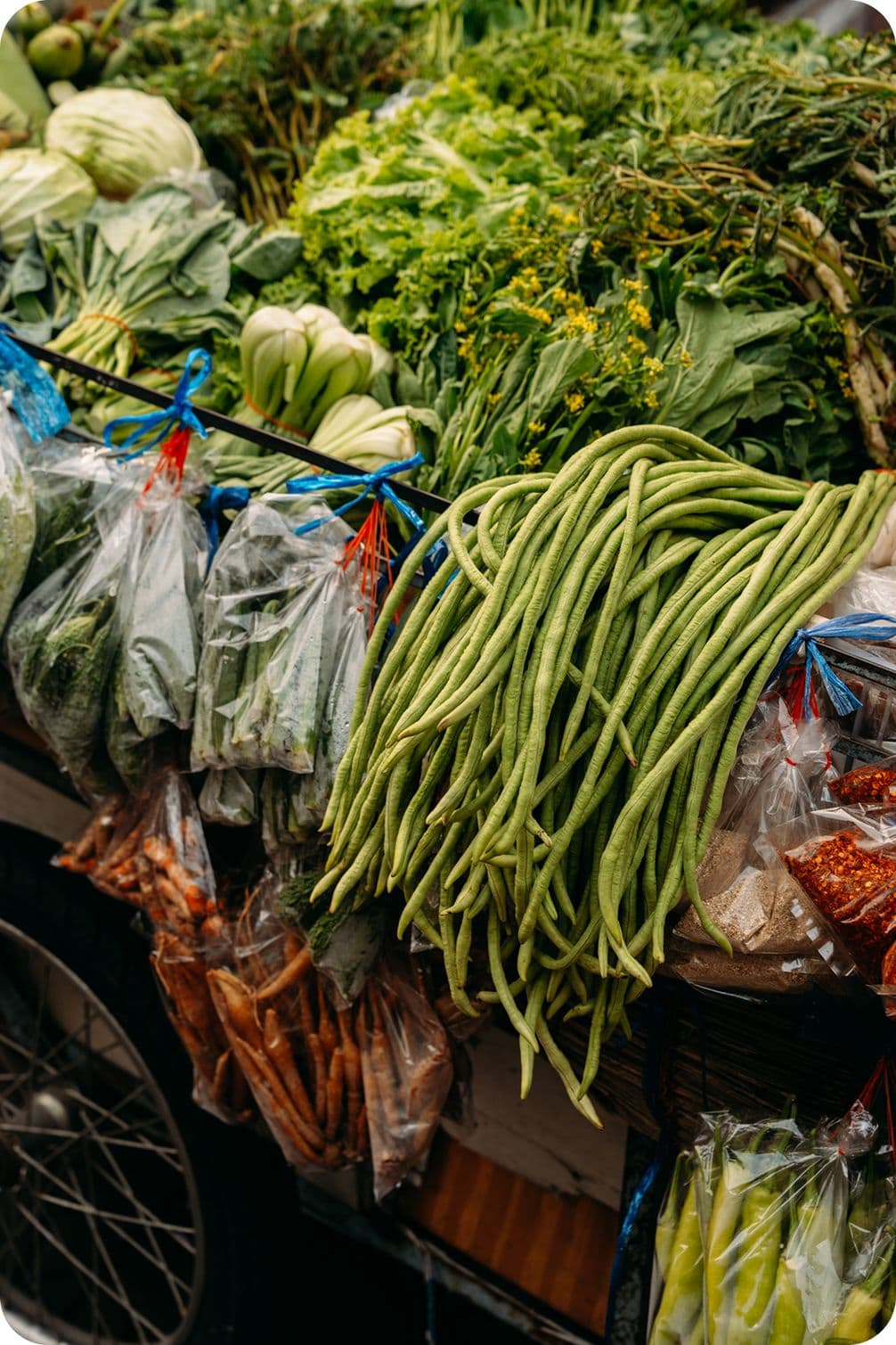 Market stall piled with long green yardlong beans, leafy greens, cabbages and plastic-bagged vegetables.