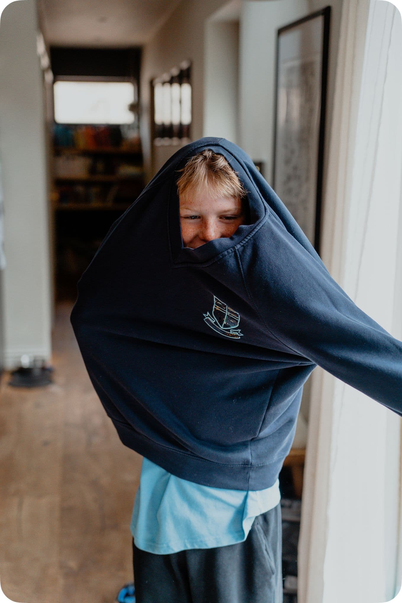 Child playfully wearing a large navy sweatshirt, arms inside, smiling in a brightly lit room with wooden floors and framed artwork.