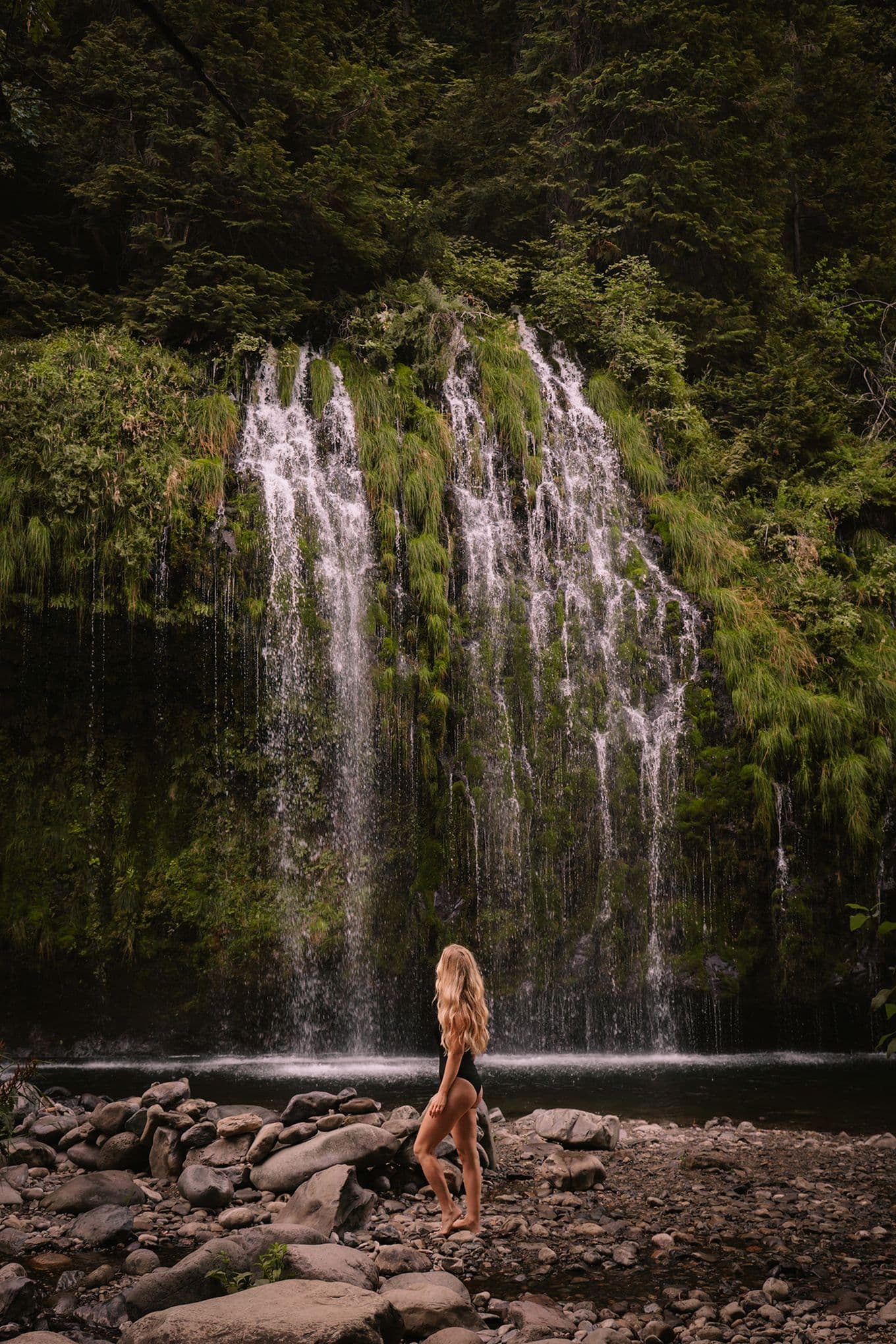 Sophie, with long blonde hairs, stands on rocks, gazing at a lush, green waterfall surrounded by dense forest.