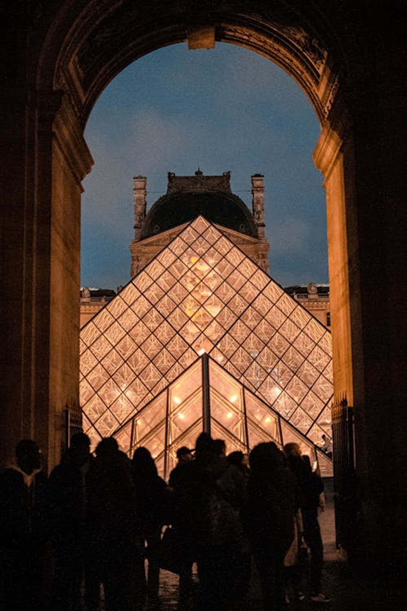 Silhouetted people stand under an archway, facing the illuminated glass pyramid of the Louvre Museum at dusk.