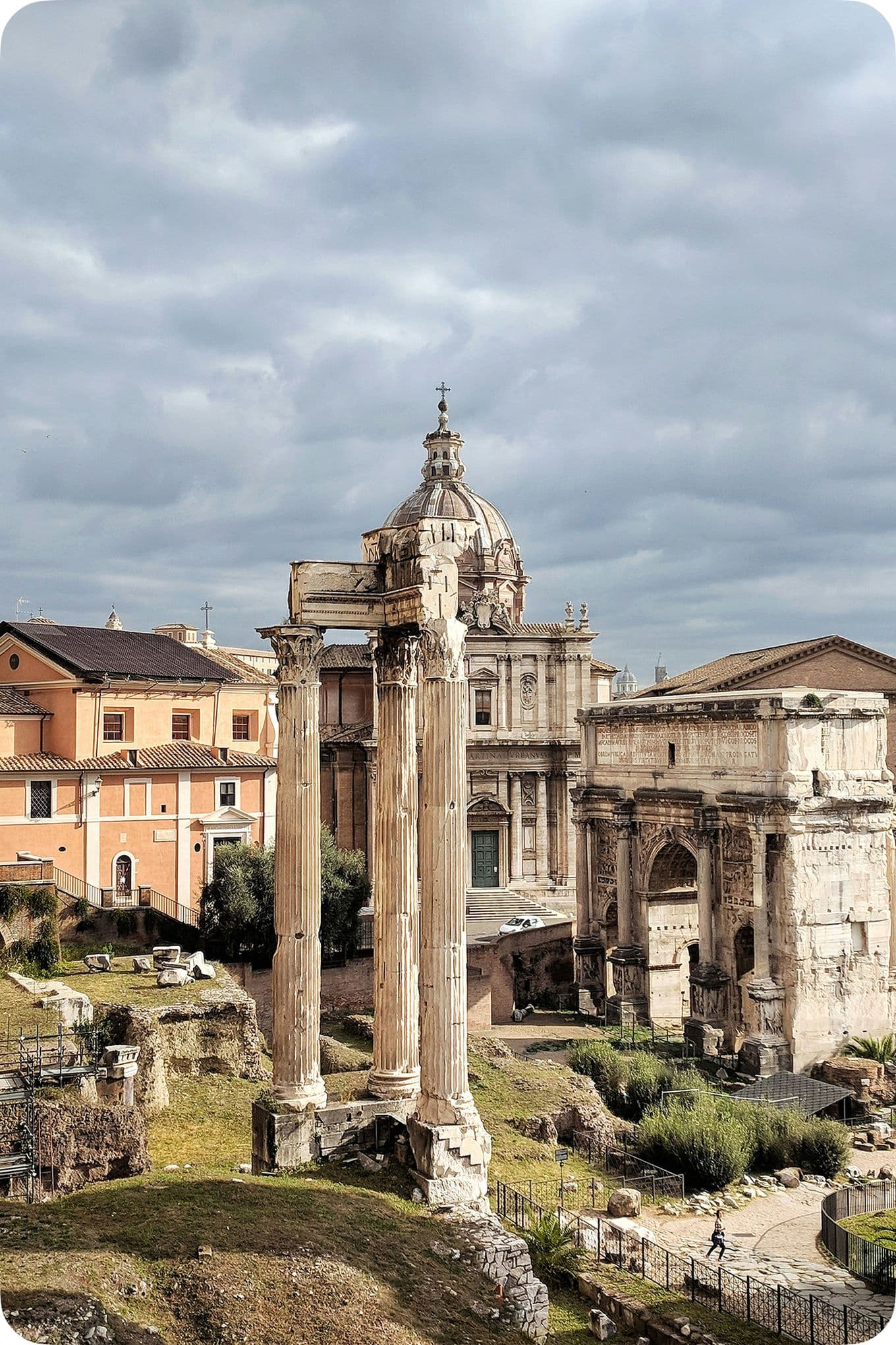 Ancient Roman ruins with three tall columns, surrounded by historic buildings under a cloudy sky.