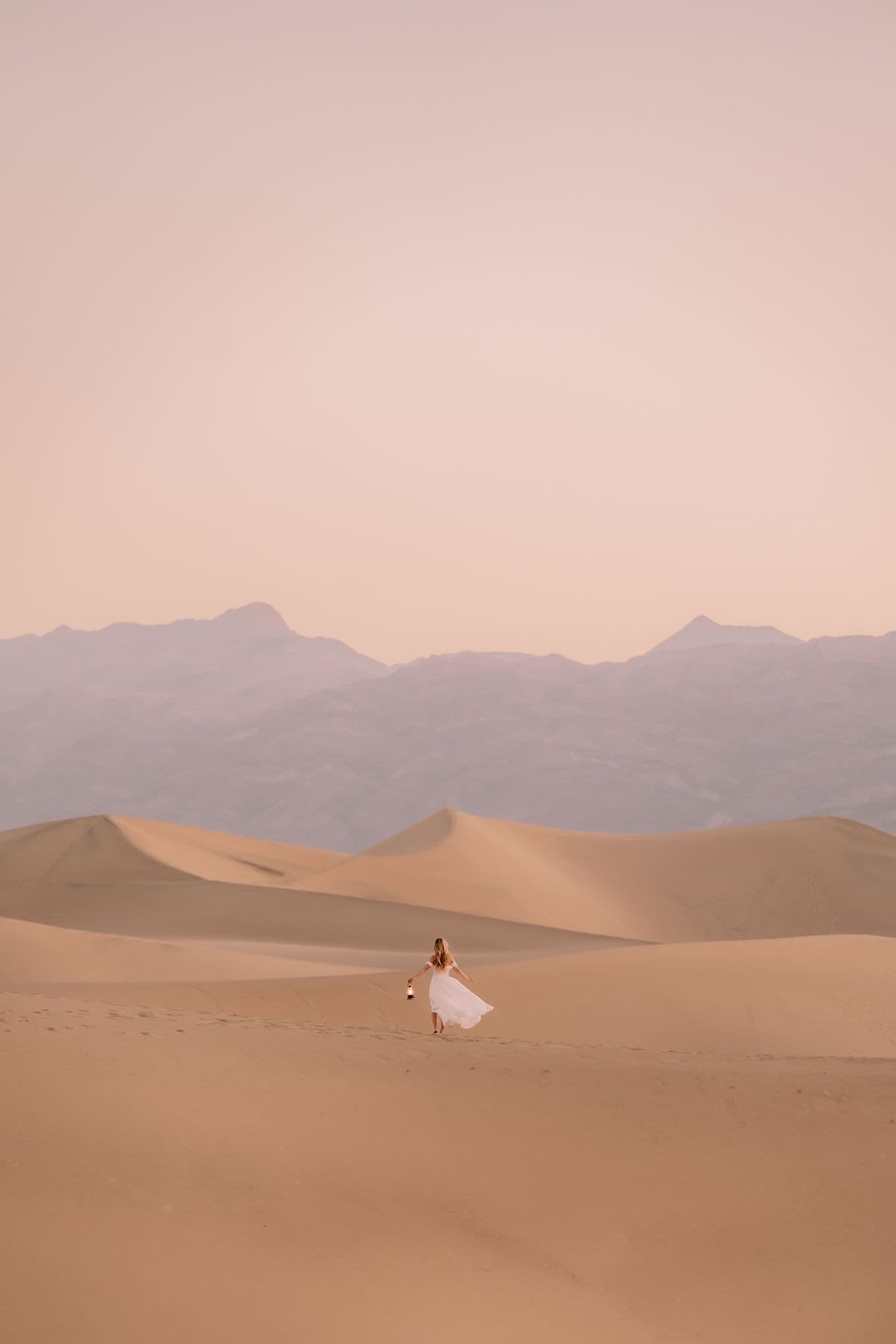 Sophie, wearing a flowing white dress, walks across vast sand dunes at sunset, with distant mountains under a soft pink sky.