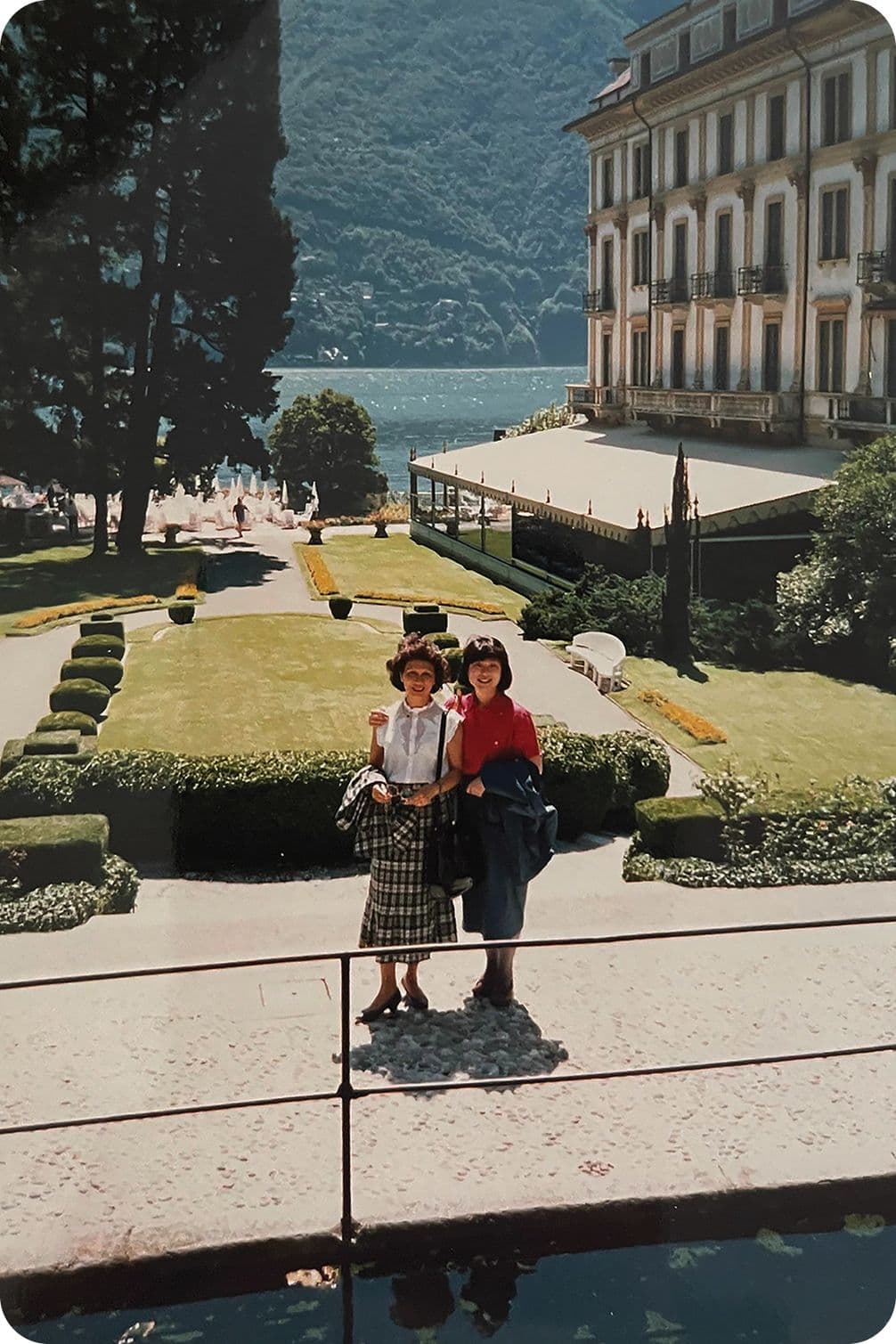 Two people posing in front of a grand building with manicured gardens, overlooking a lake and mountains under a clear sky.
