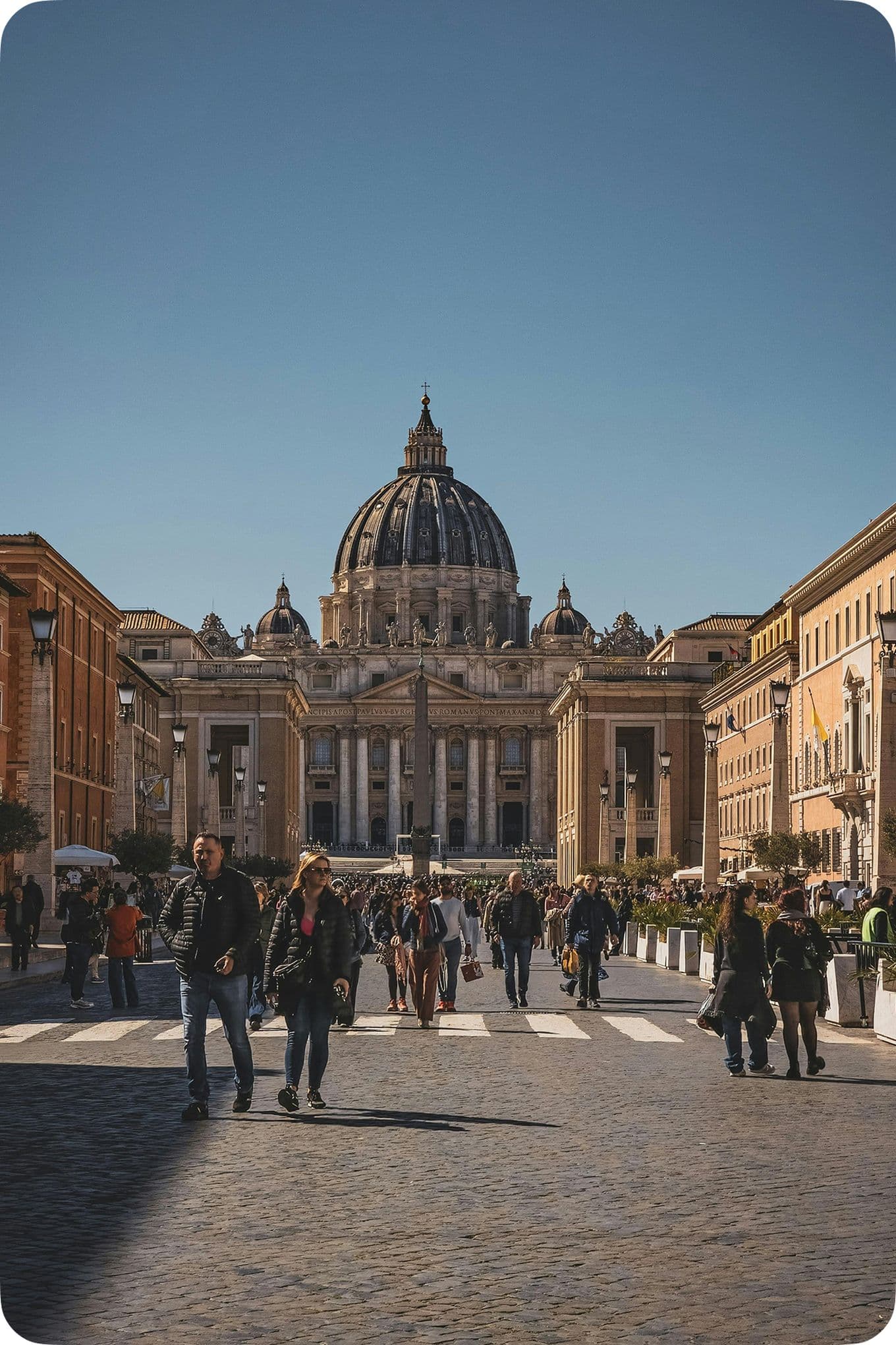 People walking towards St. Peter's Basilica in Vatican City under a clear blue sky. The street is bustling with activity.