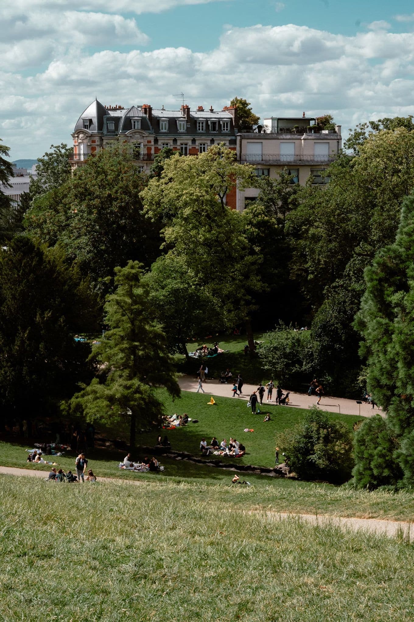 People relaxing on a grassy park hillside with trees and a historic building in the background under a cloudy sky.