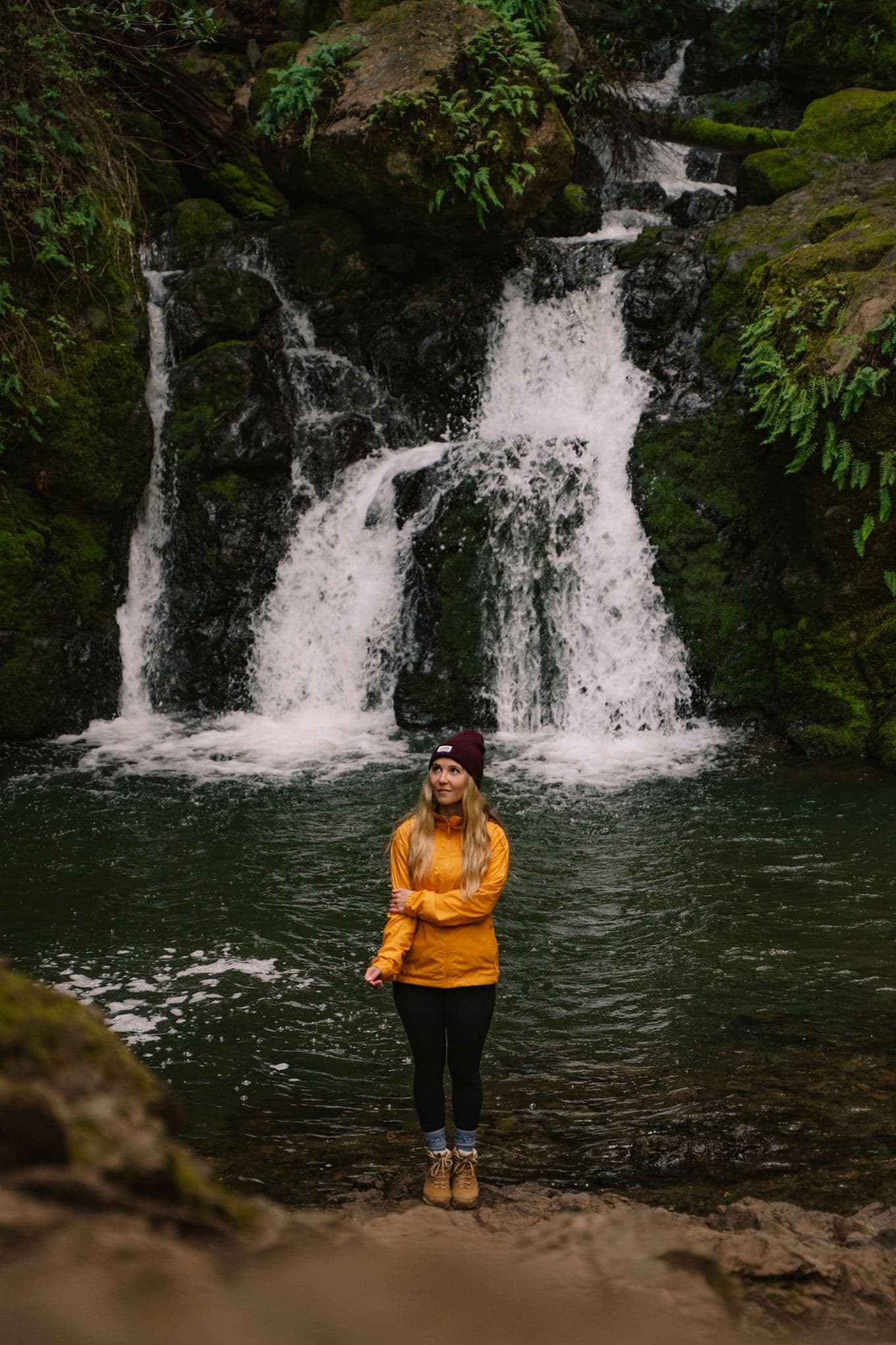 Sophie wears a yellow waterproof jacket and maroon beanie as she stands on the rocks in front of a small waterfall surrounded by green foliage