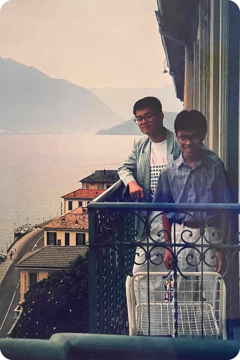 Two people stand on a balcony overlooking a scenic view of a lake and mountains, with buildings and a road below.