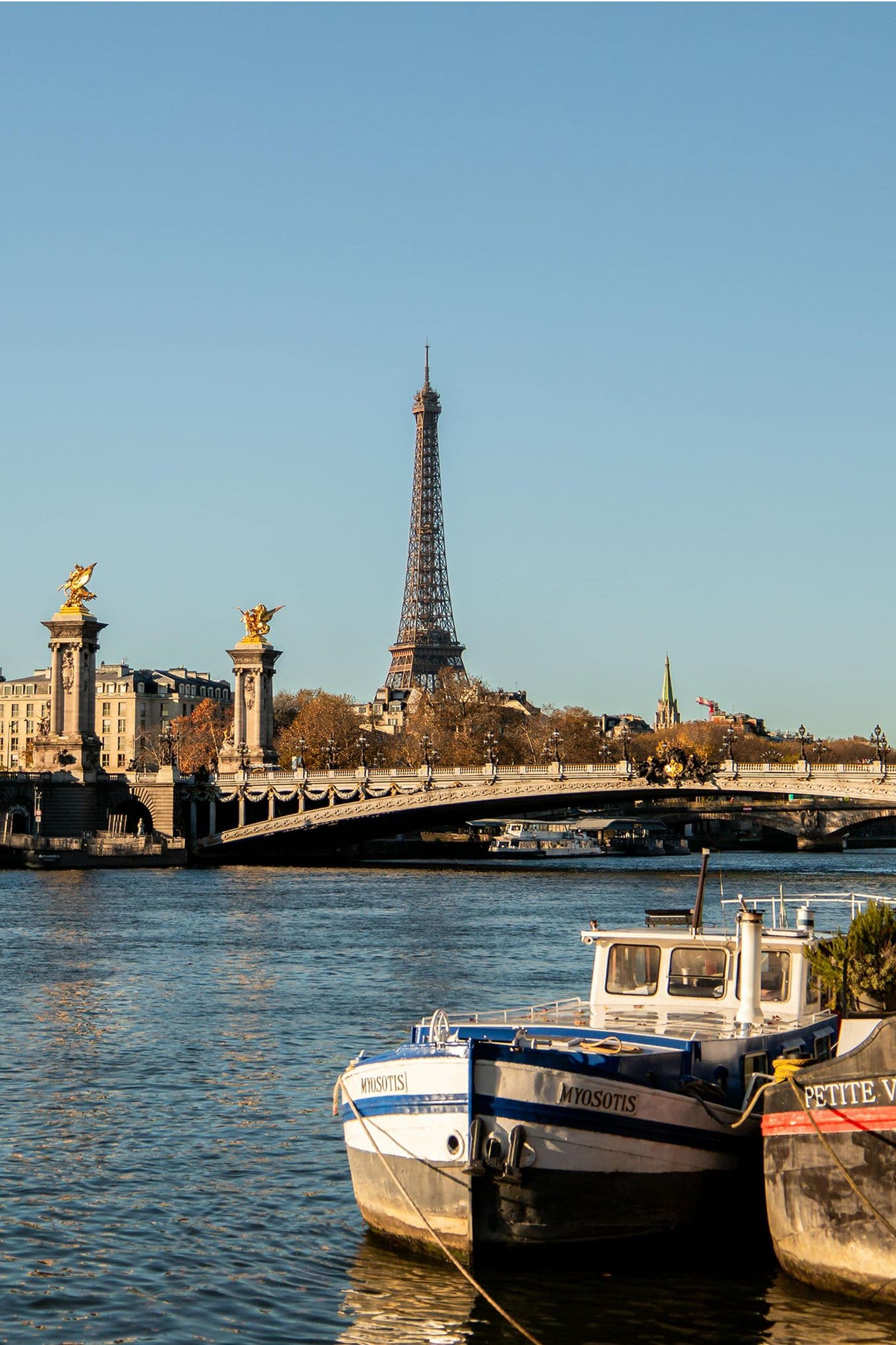 Boats docked on the Seine River with the Eiffel Tower and Pont Alexandre III bridge in the background under a clear blue sky.