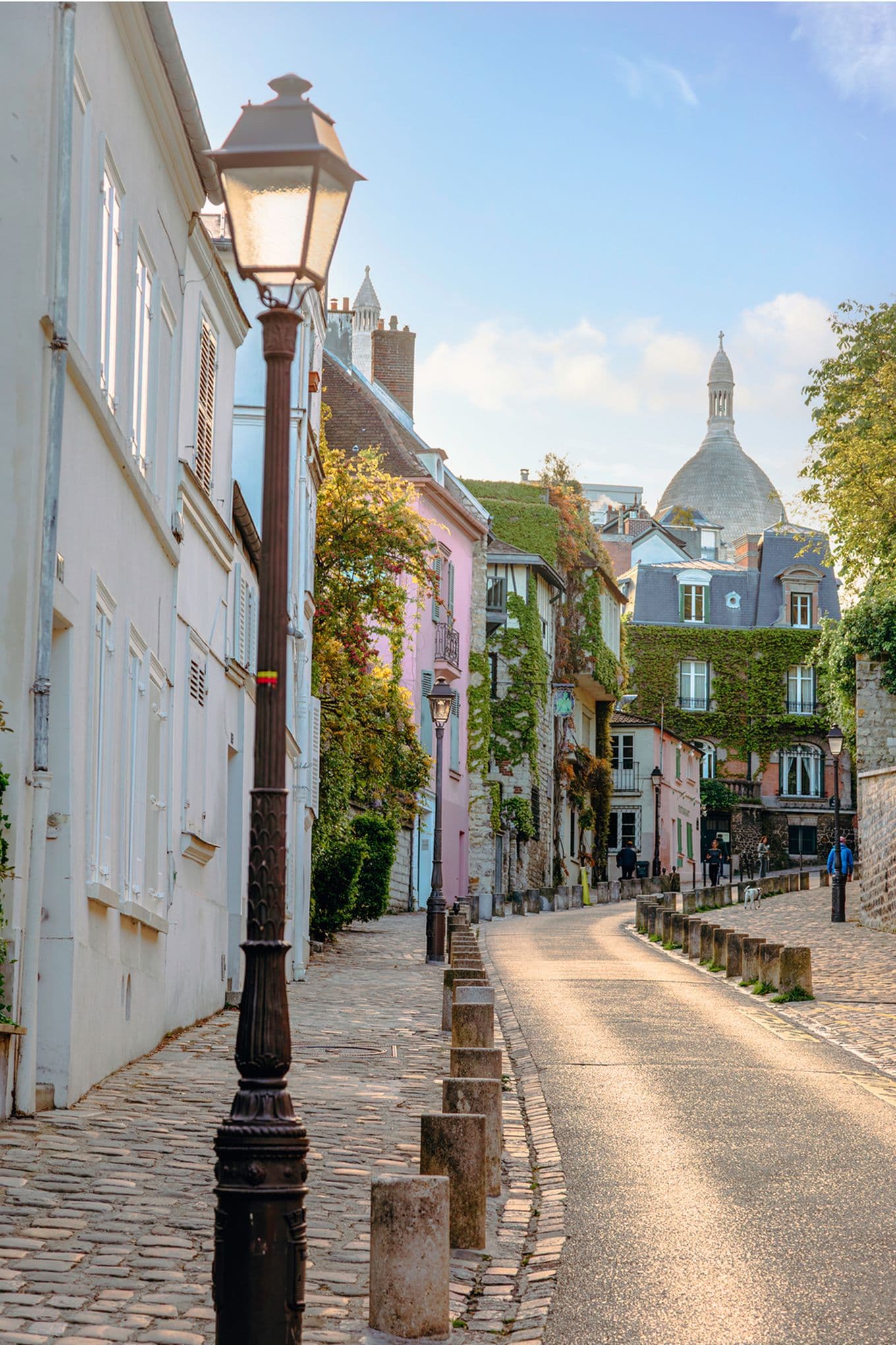 A quaint cobblestone street lined with pastel-colored buildings and vintage street lamps, leading towards a domed structure under a clear sky.