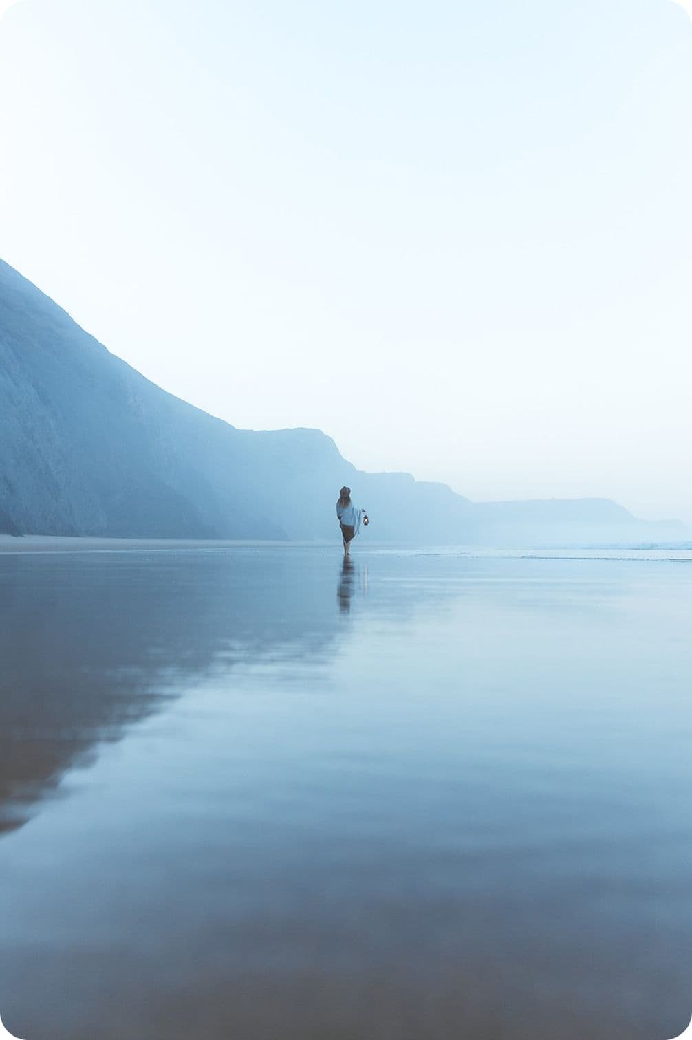 Person standing on a serene, reflective beach with misty cliffs in the background, holding a surfboard under a pale blue sky.