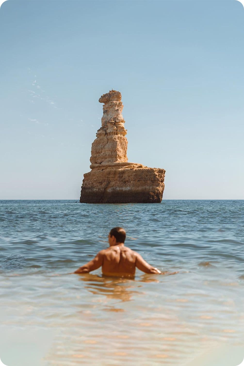 A person in the sea looks at a large, isolated rock formation under a clear blue sky.