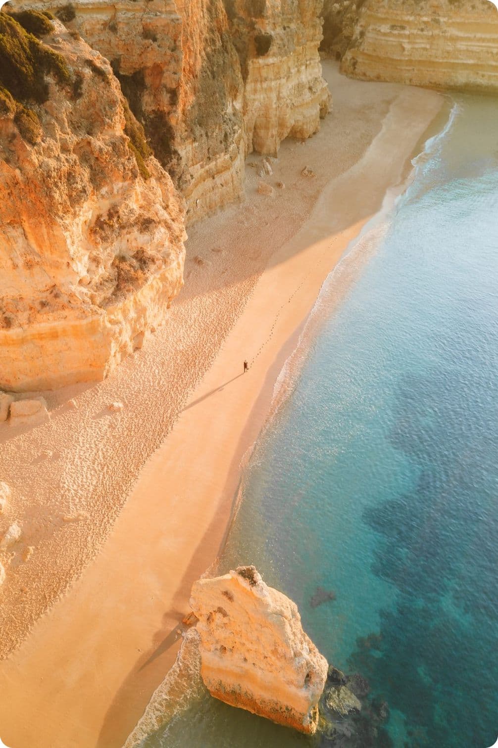 Aerial view of a sandy beach with clear turquoise water, bordered by rugged cliffs. A lone person walks along the shoreline.