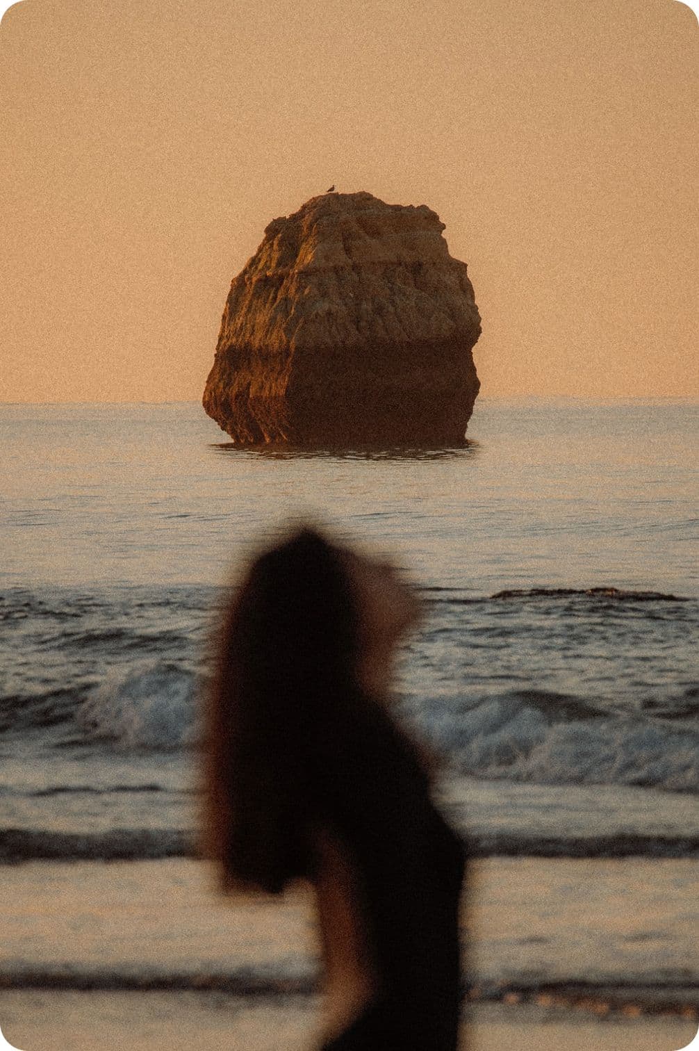 A person stands on the beach at sunset, with waves in the background and a large rock formation in the sea.