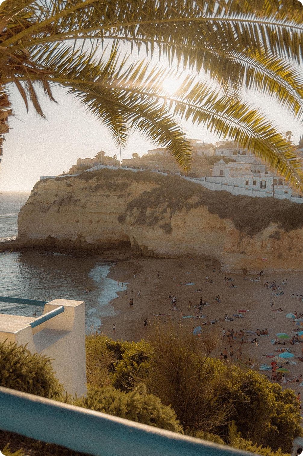 Beach scene with people on sandy shore, surrounded by cliffs and buildings, under a clear sky. Palm fronds frame the sunlit view.