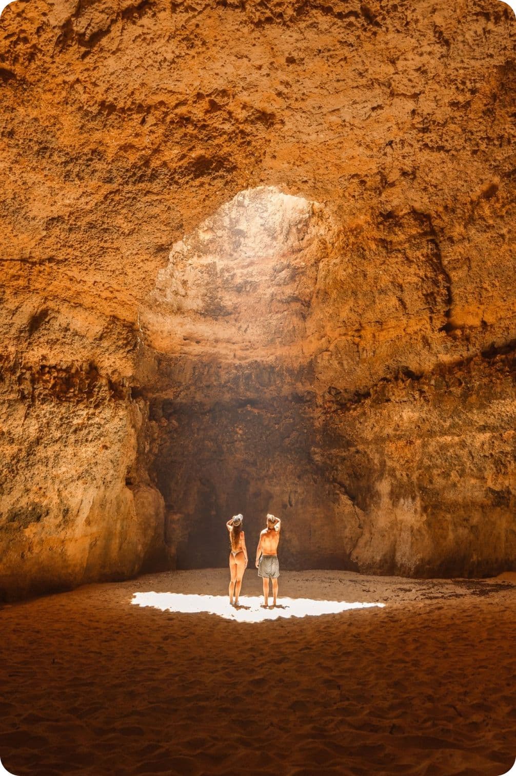Two people stand in a sunlit cave, surrounded by towering rocky walls, with light streaming through an overhead opening.