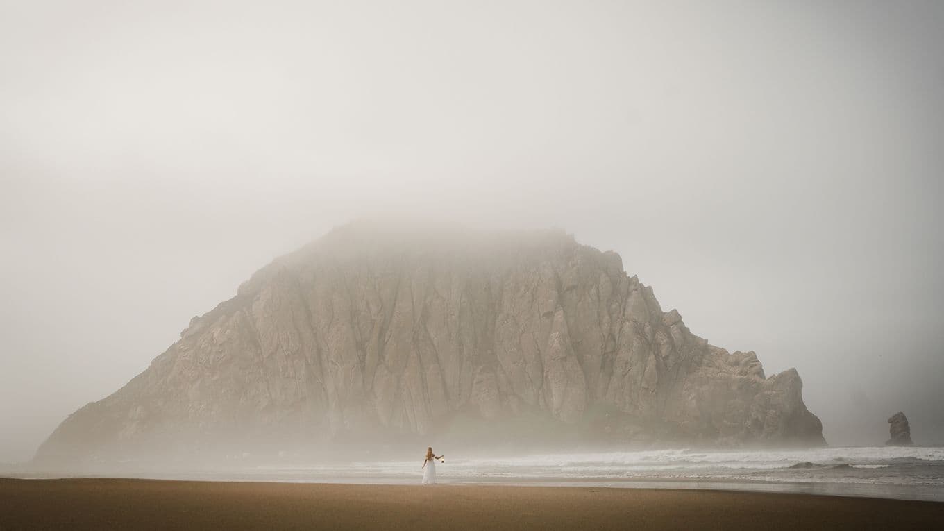A person stands on a foggy beach near a large, mist-covered rock formation, with waves gently lapping at the shore.