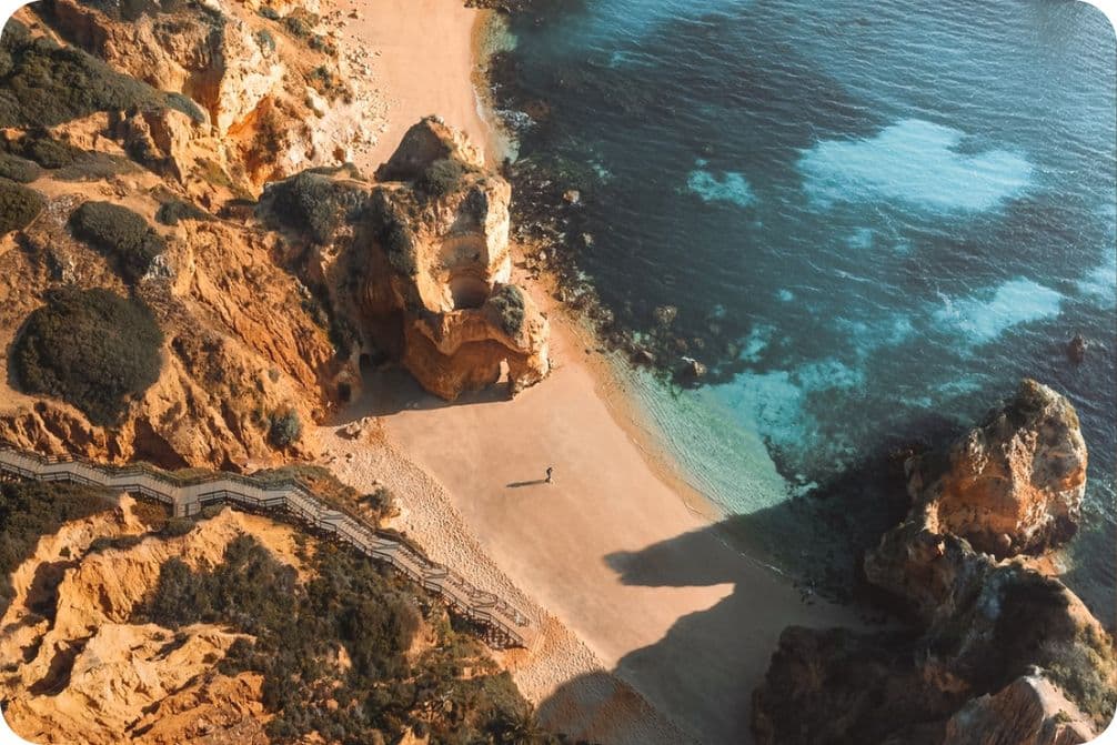 Aerial view of a secluded beach with turquoise water, surrounded by rocky cliffs and a winding wooden staircase leading down to the sand.