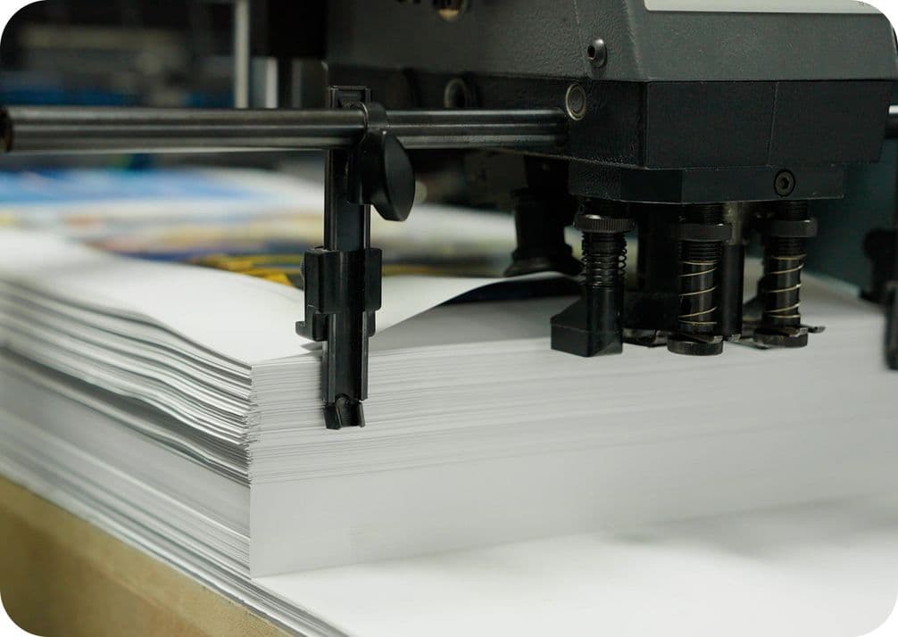 A printing machine processing a stack of white paper, with mechanical components visible, in a print shop setting.