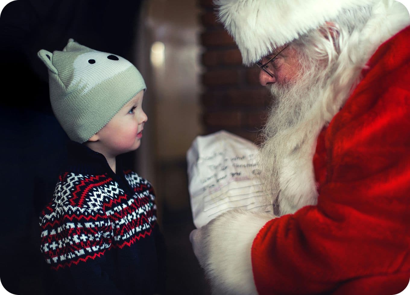 Child in a winter hat and sweater looks at Santa Claus, who is holding a letter.