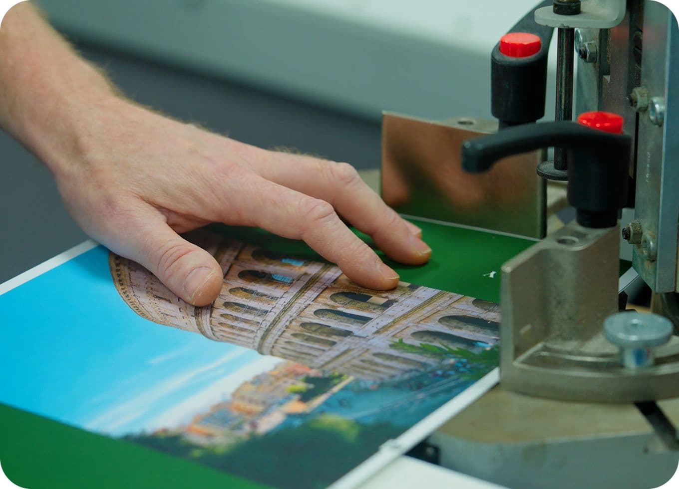 A person uses a machine to cut a laminated photo of the Colosseum, with a green background, in a print shop setting.