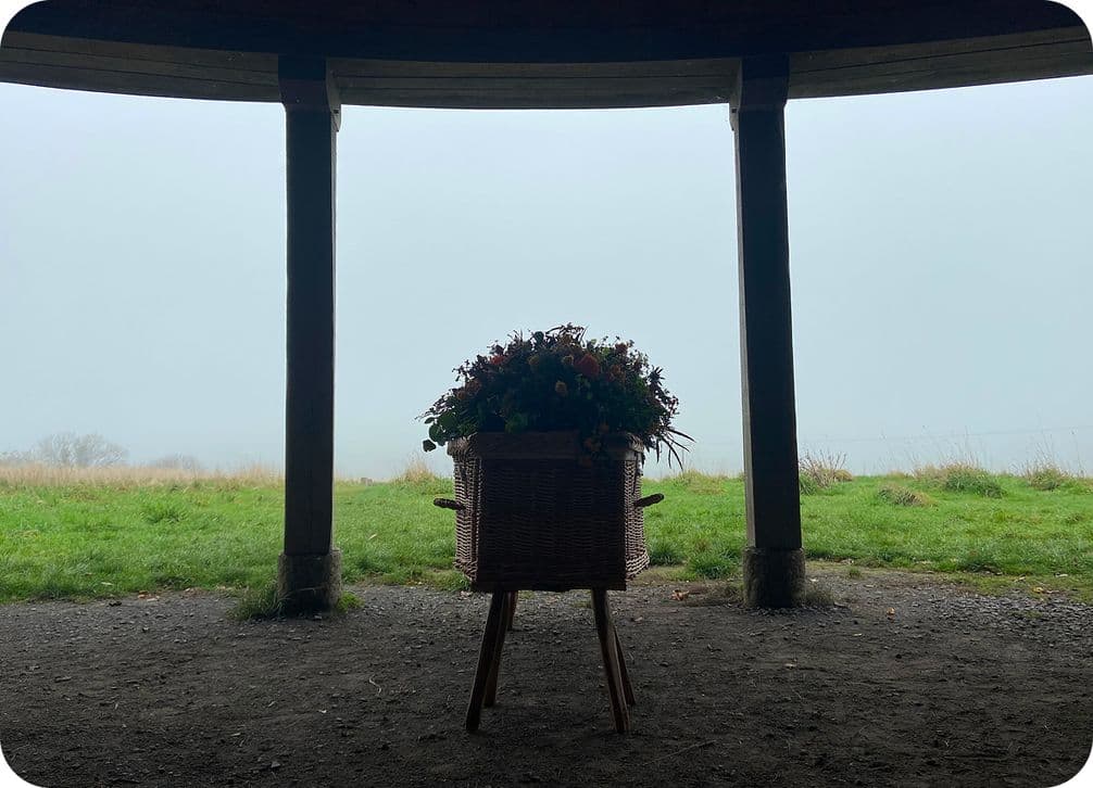 Wicker flower basket on a stand silhouetted under a pavilion, framed by wooden pillars, overlooking a foggy grassy landscape.