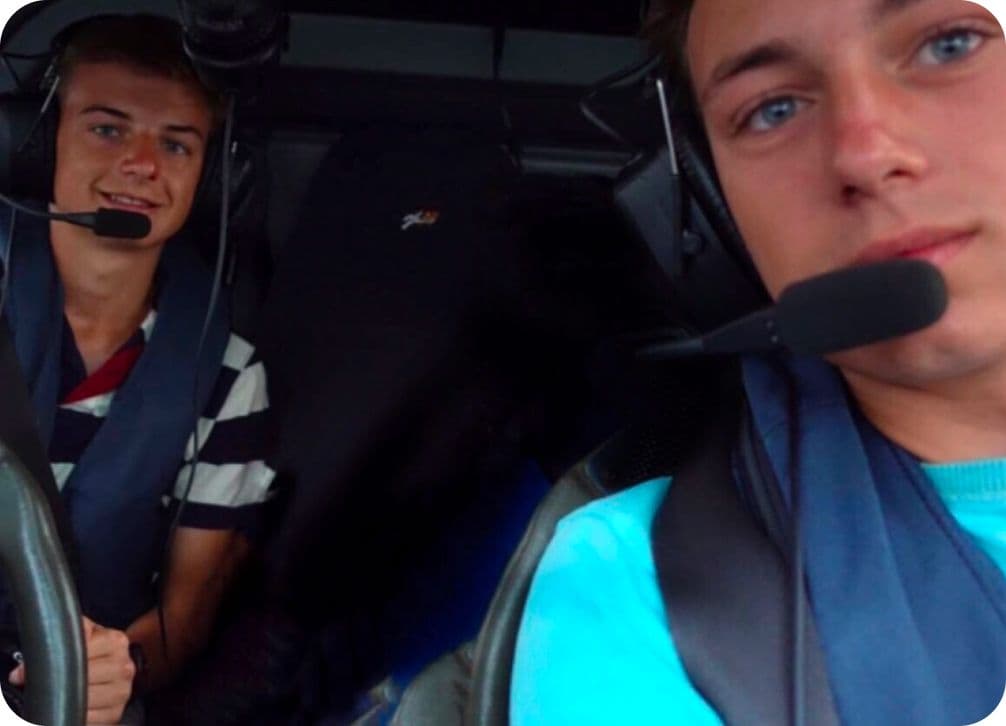 Two young men wearing headsets are seated in a vehicle, smiling at the camera.