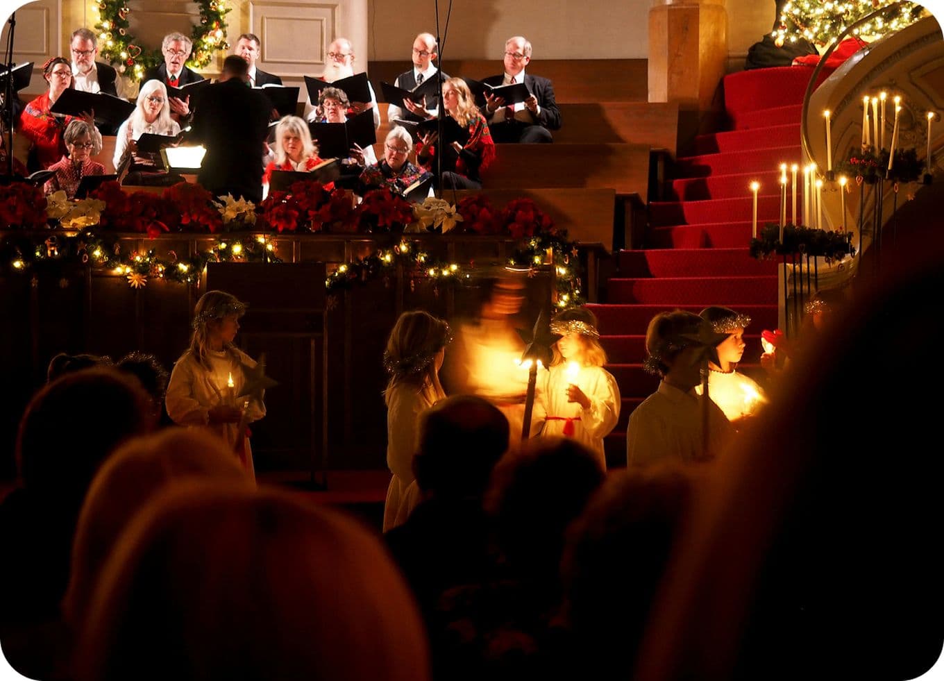 A choir performs on stage with Christmas decorations, while people holding candles walk in the foreground.