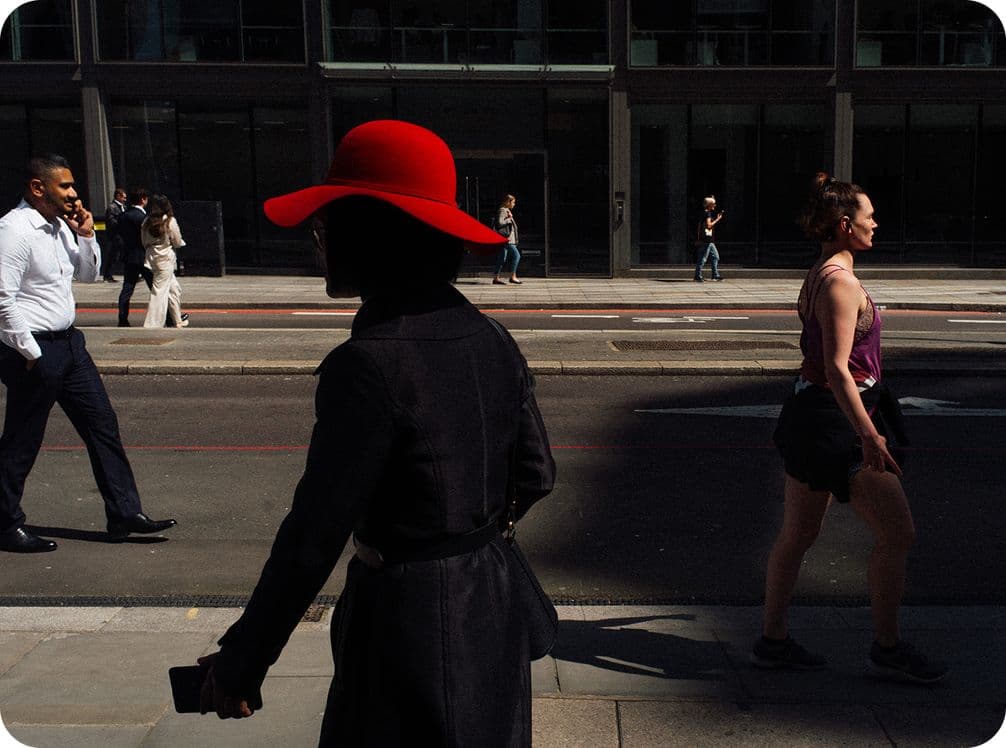 Silhouette of a person in a red hat walking on a city street, with pedestrians and a modern building in the background.
