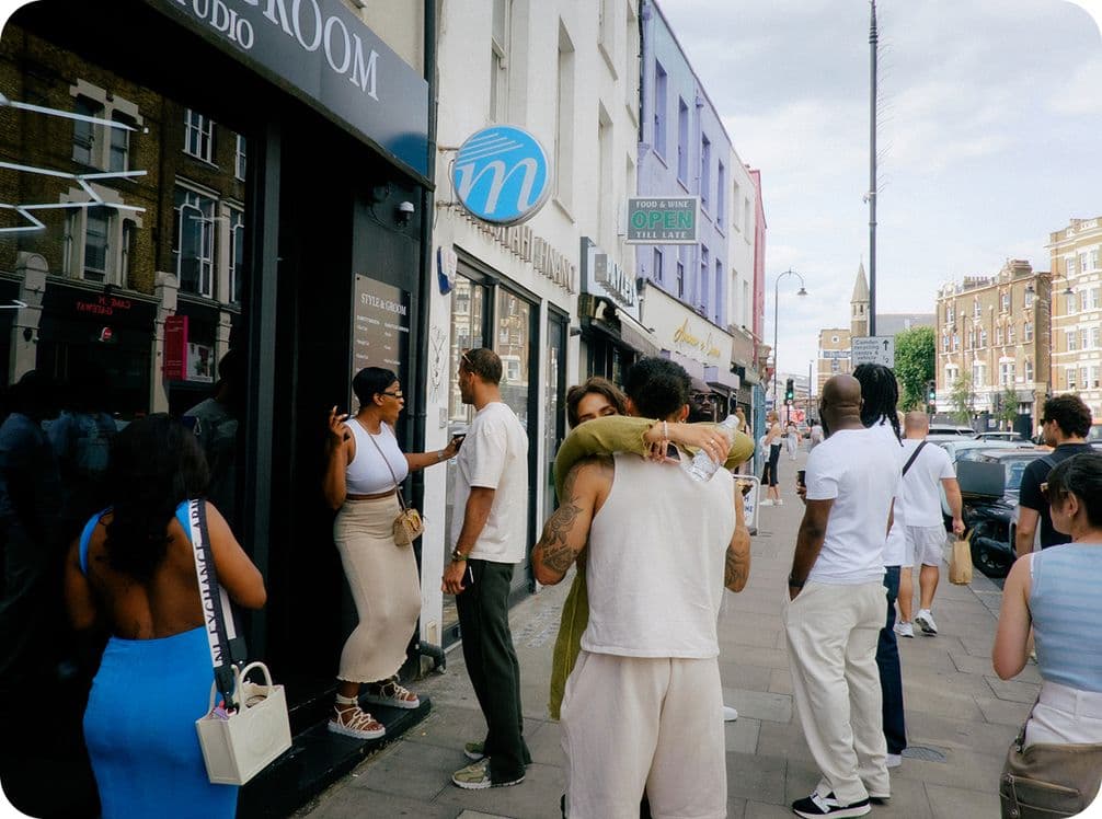 People socializing on a city street; a couple hugs, others chat outside shops. Bright day, casual attire, urban setting.