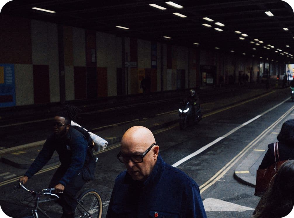 People walking and cycling in a dimly lit urban underpass, with fluorescent lights overhead and a motorcycle in the background.