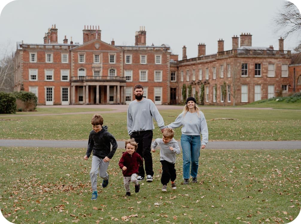 Family of five walking on grass in front of a large historic brick building with chimneys, surrounded by a park-like setting.