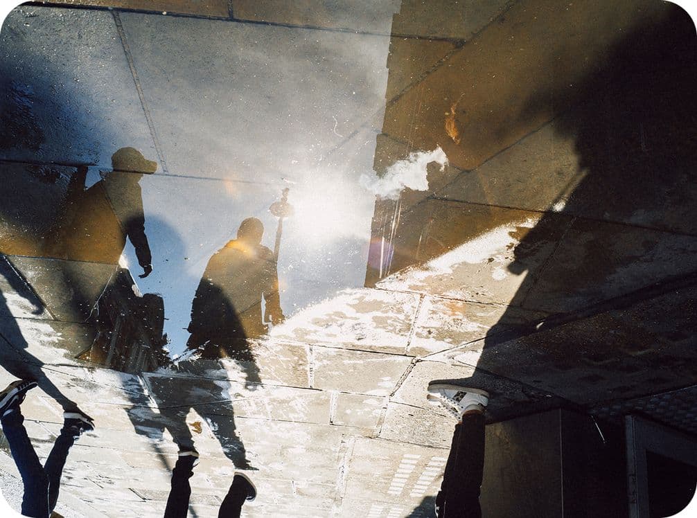Silhouettes of people and smoke reflected in a puddle on a tiled street, with sunlight creating a bright glare.