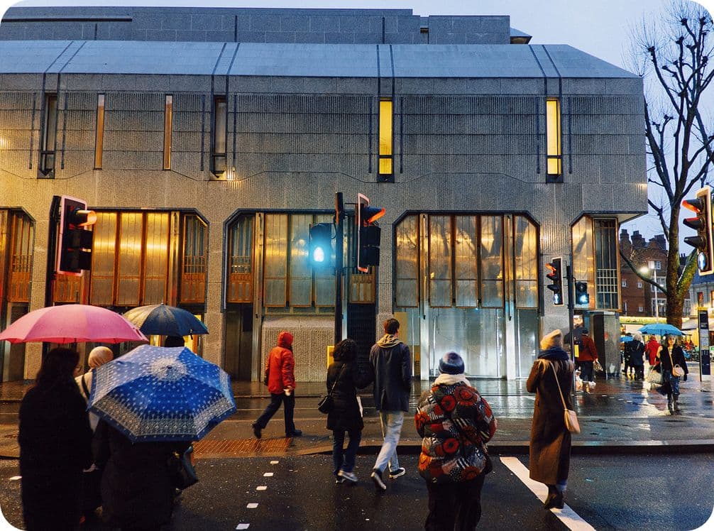 People with umbrellas crossing a street in the rain, in front of a modern gray building with tall windows and warm interior lights.